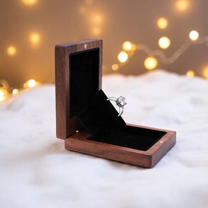 Può includere: Un anello di fidanzamento in argento con una grande gemma trasparente è esposto in un portagioie di legno aperto. La scatola è foderata in velluto nero e poggia su una superficie bianca e soffice. Luci calde sfocate sullo sfondo.