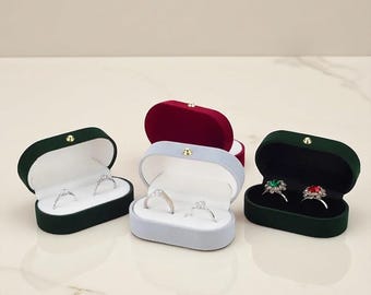 Puede incluir: Varias cajas de anillos de terciopelo en varios colores, incluyendo verde, burdeos y azul claro, están abiertas para mostrar anillos. Las cajas tienen un diseño con bisagras y un interior blanco contrastante. Algunos anillos tienen piedras transparentes, otros piedras verdes o rojas.