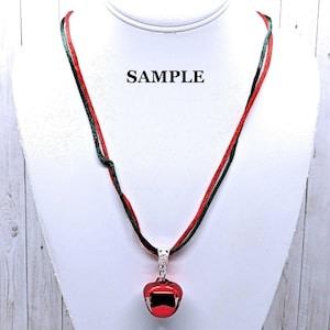 May include: A red and green ribbon necklace with a silver bell charm. The bell is red and has a shiny finish. The necklace is on a white mannequin bust. The word "SAMPLE" is written on the bust.