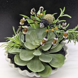 May include: A decorative spider ornament crafted from black, brown, and amber-colored beads with silver-toned accents. The spider features a black crystal-encrusted head and a textured, pale green spherical abdomen, perched atop a green succulent plant.