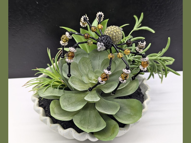 May include: A decorative spider ornament crafted from black, brown, and amber-colored beads with silver-toned accents. The spider features a black crystal-encrusted head and a textured, pale green spherical abdomen, perched atop a green succulent plant.