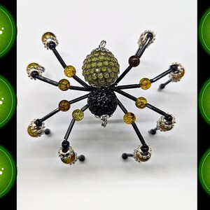 May include: A spider-shaped ornament featuring a large, textured olive-green bead for the head and a smaller, black, faceted bead for the abdomen. Eight black wire legs are adorned with smaller amber-colored crackle beads and silver-toned accents.