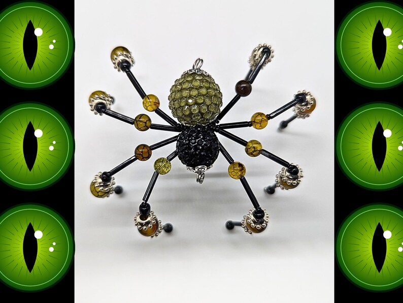 May include: A spider-shaped ornament featuring a large, textured olive-green bead for the head and a smaller, black, faceted bead for the abdomen. Eight black wire legs are adorned with smaller amber-colored crackle beads and silver-toned accents.