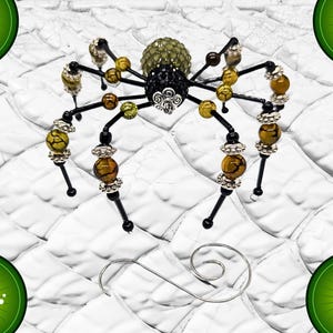 May include: A decorative spider ornament crafted from black wire, featuring a textured olive green abdomen and a black crystal-encrusted thorax. The eight legs are adorned with yellow and brown crackle beads and silver-toned metal spacers.