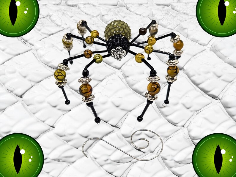 May include: A decorative spider ornament crafted from black wire, featuring a textured olive green abdomen and a black crystal-encrusted thorax. The eight legs are adorned with yellow and brown crackle beads and silver-toned metal spacers.