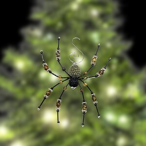 May include: A decorative spider ornament featuring a textured olive green abdomen and a black head. The eight legs are constructed from thin black wire accented with brown beads and silver-toned spacers. A silver metal hook is attached to the top.