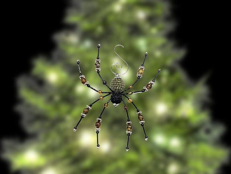 May include: A decorative spider ornament featuring a textured olive green abdomen and a black head. The eight legs are constructed from thin black wire accented with brown beads and silver-toned spacers. A silver metal hook is attached to the top.
