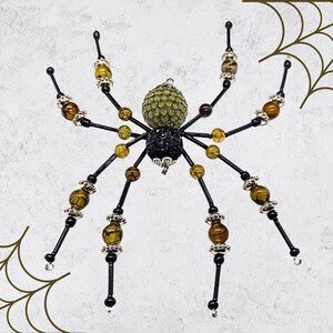 May include: A decorative spider ornament featuring a black and olive green beaded body. The eight legs are constructed from black wire accented with amber-colored beads, clear faceted beads, and silver-toned metal spacers throughout the design.