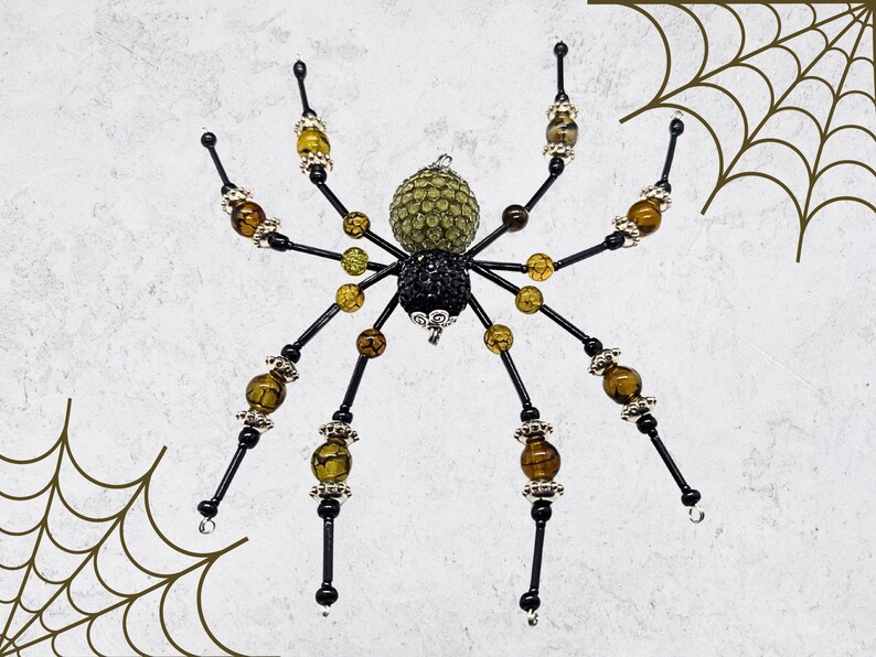 May include: A decorative spider ornament featuring a black and olive green beaded body. The eight legs are constructed from black wire accented with amber-colored beads, clear faceted beads, and silver-toned metal spacers throughout the design.