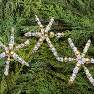 May include: Three white and gold beaded snowflake ornaments on a green evergreen background.
