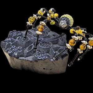 May include: A decorative spider figurine crafted from black wire and beads, featuring amber-colored and yellow crackle glass beads on its legs, a black and silver patterned head, and a textured yellow body, perched on a dark carved wooden block.