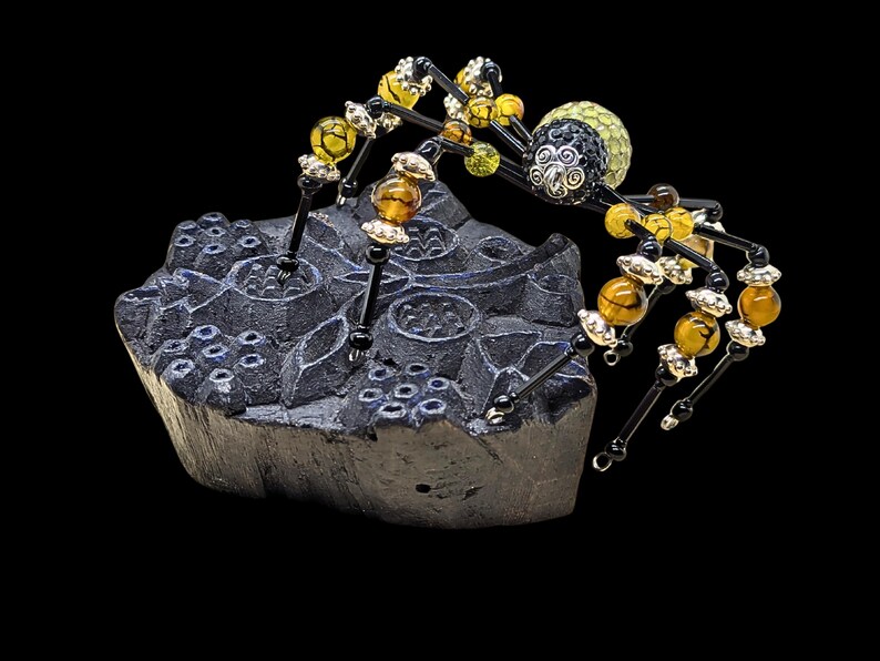 May include: A decorative spider figurine crafted from black wire and beads, featuring amber-colored and yellow crackle glass beads on its legs, a black and silver patterned head, and a textured yellow body, perched on a dark carved wooden block.