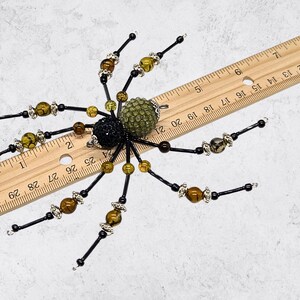 May include: A decorative spider ornament crafted from black, olive green, and brown beads with silver-toned accents. The spider is positioned over a wooden ruler, showing a total width of approximately 5 inches from leg tip to leg tip.