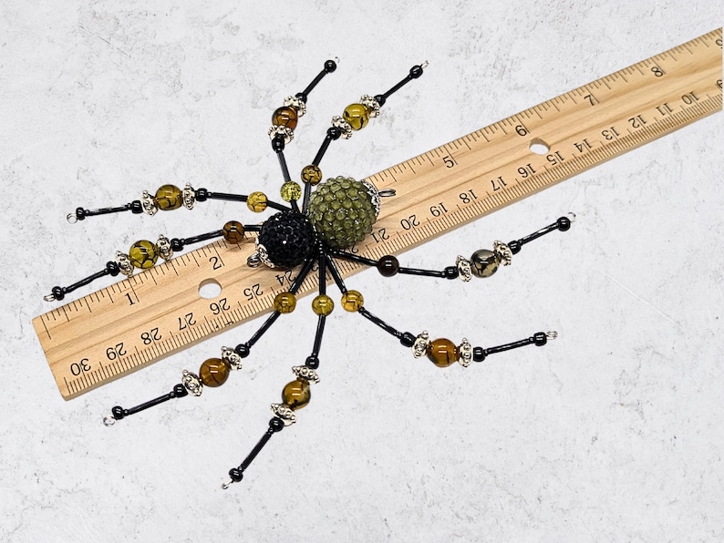 May include: A decorative spider ornament crafted from black, olive green, and brown beads with silver-toned accents. The spider is positioned over a wooden ruler, showing a total width of approximately 5 inches from leg tip to leg tip.