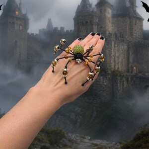 May include: A spider-shaped ring featuring a textured green bead for the abdomen, a black central body, and eight spindly black legs accented with small amber-colored beads and silver-toned metal spacers, worn across the back of the hand.