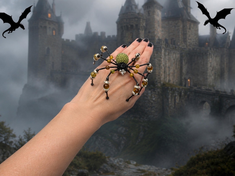 May include: A spider-shaped ring featuring a textured green bead for the abdomen, a black central body, and eight spindly black legs accented with small amber-colored beads and silver-toned metal spacers, worn across the back of the hand.