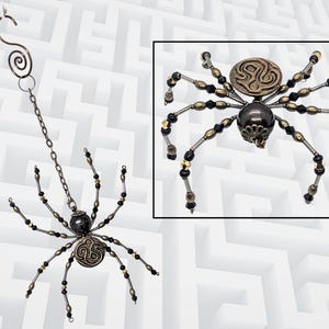 May include: A decorative spider ornament featuring a dark metallic body and a textured bronze-toned circular back. The eight legs are crafted from wire strung with alternating black and metallic beads. A metal chain and spiral hook allow for hanging.