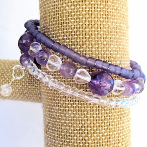 May include: A set of three bracelets featuring amethyst and clear glass beads. The bracelets are layered on a brown fabric display stand.