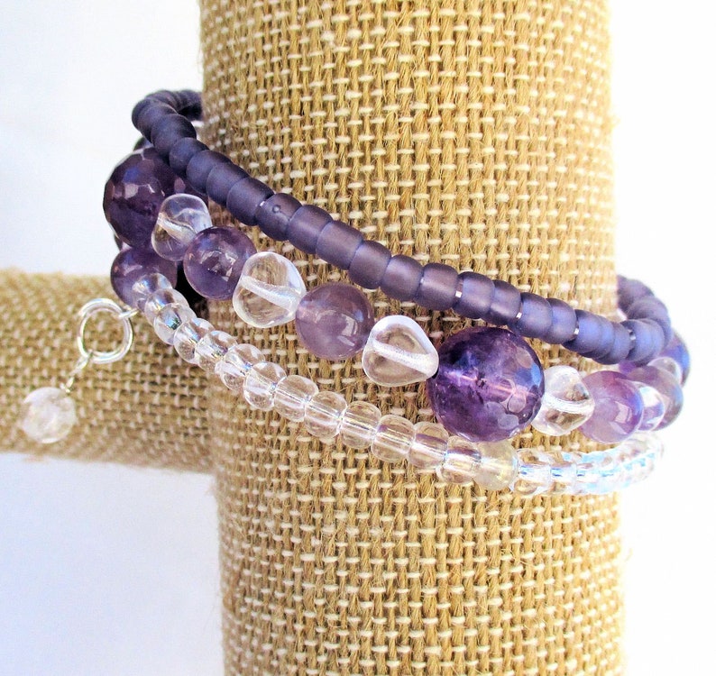 May include: A set of three bracelets featuring amethyst and clear glass beads. The bracelets are layered on a brown fabric display stand.