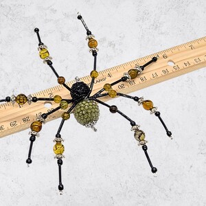 May include: A decorative spider ornament featuring a black textured head and a green beaded abdomen. The eight legs are made of black wire accented with amber-colored crackle beads and silver-toned spacers. A wooden ruler shows a width of 8 inches.