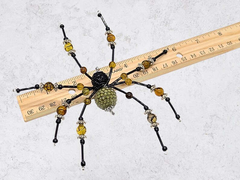 May include: A decorative spider ornament featuring a black textured head and a green beaded abdomen. The eight legs are made of black wire accented with amber-colored crackle beads and silver-toned spacers. A wooden ruler shows a width of 8 inches.