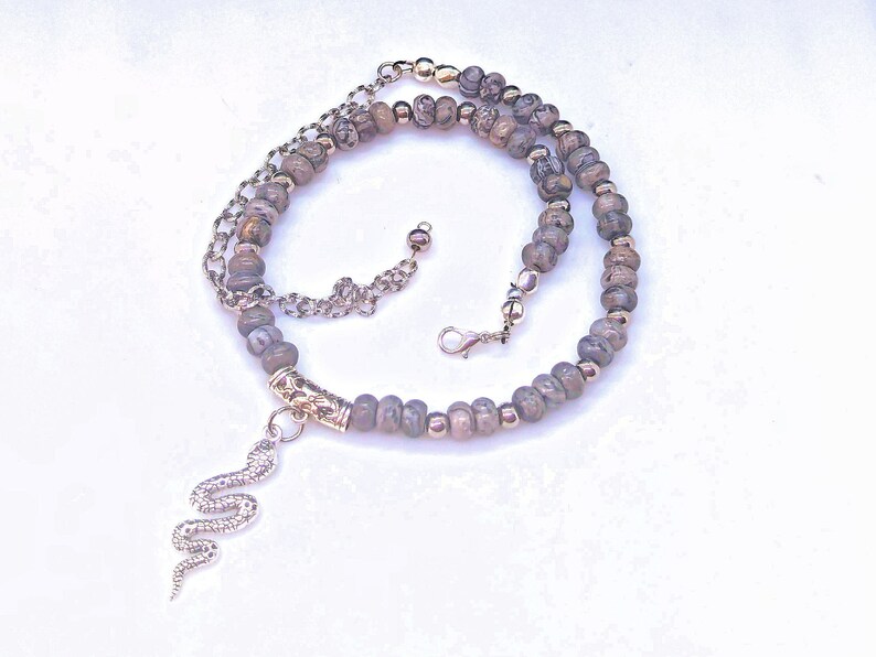 May include: A silver snake charm hangs from a layered necklace with brown and silver beads.