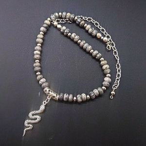 May include: A silver snake charm hangs from a grey and white stone beaded necklace with a silver chain.