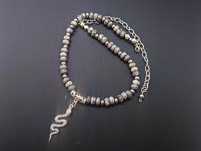 May include: A silver snake charm hangs from a grey and white stone beaded necklace with a silver chain.
