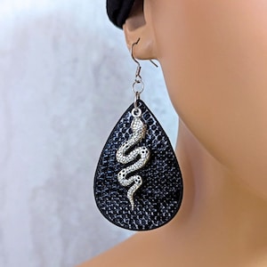 May include: A teardrop-shaped earring featuring a black textured surface with a snakeskin-like pattern. A silver-toned metal snake charm is attached to the center of the earring, which hangs from a standard silver-toned metal fishhook ear wire.