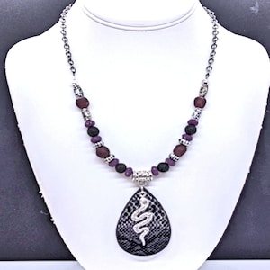 May include: A silver chain necklace with a black and silver snake pendant. The necklace is made with purple and black beads.