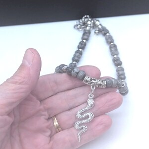 May include: A grey beaded necklace with a silver snake pendant. The necklace is made of round beads and has a silver clasp.