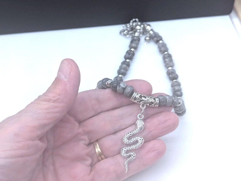 May include: A grey beaded necklace with a silver snake pendant. The necklace is made of round beads and has a silver clasp.