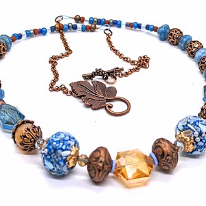 May include: A copper and blue beaded necklace with a leaf-shaped clasp. The necklace features a variety of blue and copper beads, including some with intricate designs.