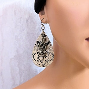 May include: A pair of dangle earrings with a teardrop-shaped design. The earrings are made of white faux leather with a black snake design. The snake is coiled and has a gold finish.