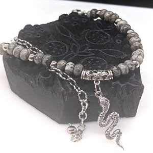 May include: A grey and silver beaded bracelet with a snake charm. The bracelet is made with grey and white stones and has a silver clasp. The snake charm is silver and has a detailed design.