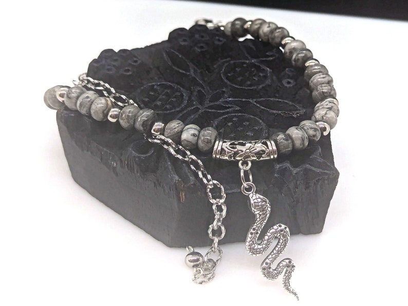 May include: A grey and silver beaded bracelet with a snake charm. The bracelet is made with grey and white stones and has a silver clasp. The snake charm is silver and has a detailed design.