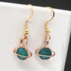 May include: A pair of gold earrings with a green planet charm. The planet has a gold ring around it.