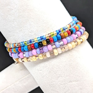 May include: A stack of colorful beaded bracelets in shades of blue, red, pink, purple, brown, and gold. The bracelets are made of small, round beads and are layered on top of each other.
