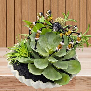 May include: A decorative spider ornament crafted from black, amber, and silver-toned beads, featuring a textured body and long legs, perched atop a cluster of artificial green succulents housed in a white, scalloped-edge ceramic bowl.