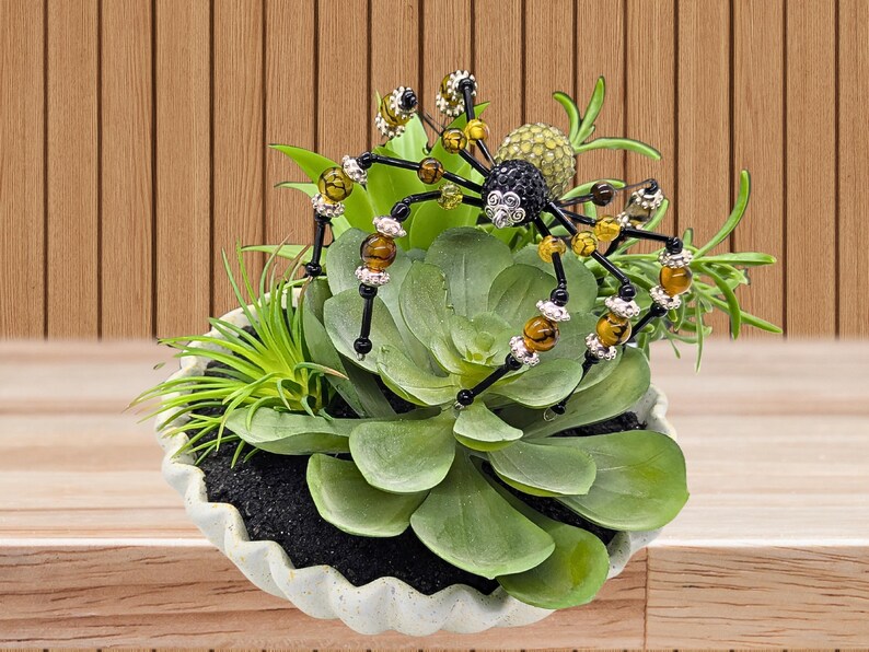 May include: A decorative spider ornament crafted from black, amber, and silver-toned beads, featuring a textured body and long legs, perched atop a cluster of artificial green succulents housed in a white, scalloped-edge ceramic bowl.