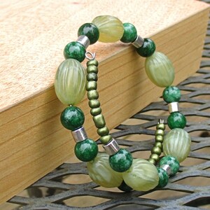 May include: A green and light green beaded bracelet with silver accents. The bracelet is made of oval-shaped beads and is wrapped around a wooden surface.