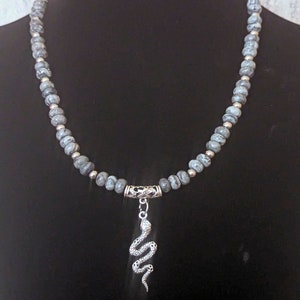 May include: A grey and silver necklace with a snake pendant. The necklace is made of small, round beads with a marbled pattern.