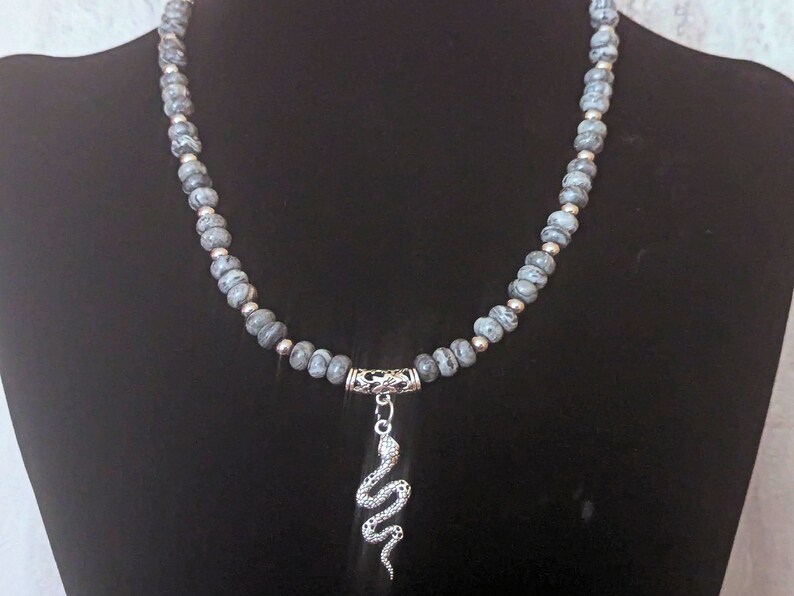 May include: A grey and silver necklace with a snake pendant. The necklace is made of small, round beads with a marbled pattern.