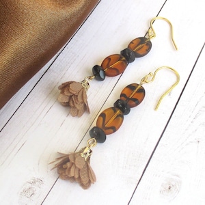 May include: A pair of dangle earrings with brown and black beads and a brown fabric tassel. The earrings have gold hooks.