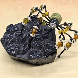 May include: A decorative spider figurine crafted from black wire, amber-colored crackle beads, and silver-toned metal spacers. The spider features a textured black head and a pale green, spherical abdomen, perched atop a carved black block.