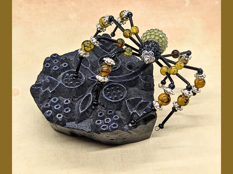 May include: A decorative spider figurine crafted from black wire, amber-colored crackle beads, and silver-toned metal spacers. The spider features a textured black head and a pale green, spherical abdomen, perched atop a carved black block.