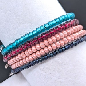 May include: Five beaded bracelets stacked on a white surface. The bracelets are teal, burgundy, light pink, dark blue, and black.