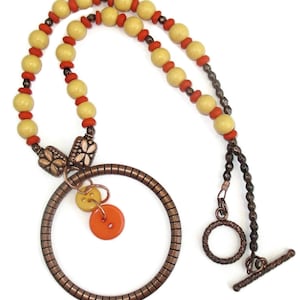 May include: A copper necklace with a circle pendant and two buttons. The necklace is made with yellow and orange beads and a copper chain.