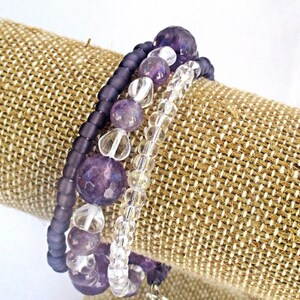 May include: A multi-strand bracelet featuring amethyst and clear glass beads. The bracelet is wrapped around a brown fabric display stand.