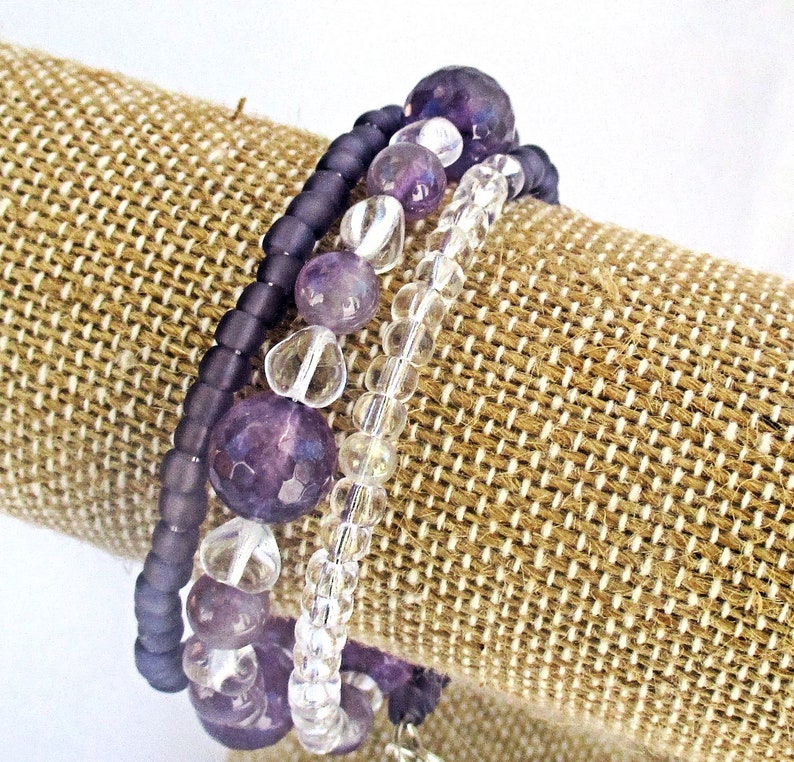 May include: A multi-strand bracelet featuring amethyst and clear glass beads. The bracelet is wrapped around a brown fabric display stand.